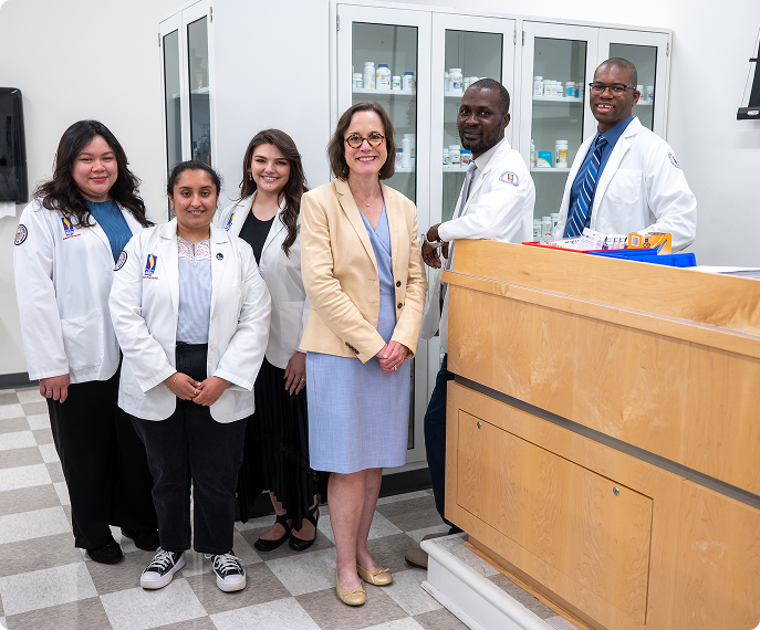 A group of pharmacy students smiles in the lab while wearing white coats. Mandy Reece stands in the center smiling in a blue dress and beige blazer.