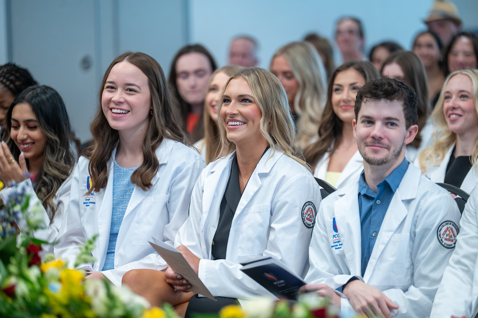 a group of students sits in the audience of the white coat ceremony and smiles as they watch the presentation