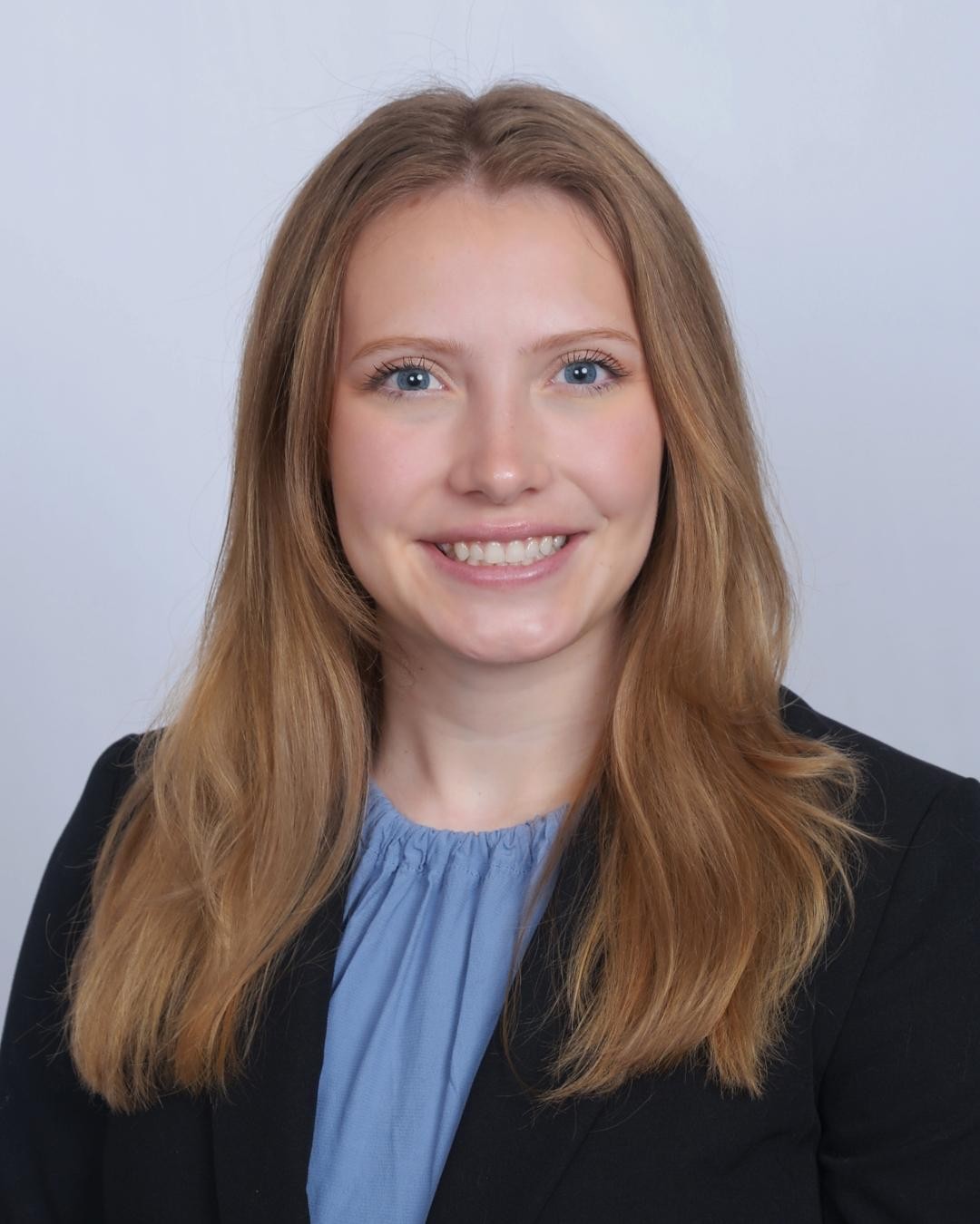 A female student smiles in a blue blouse and black blazer against a a gray background