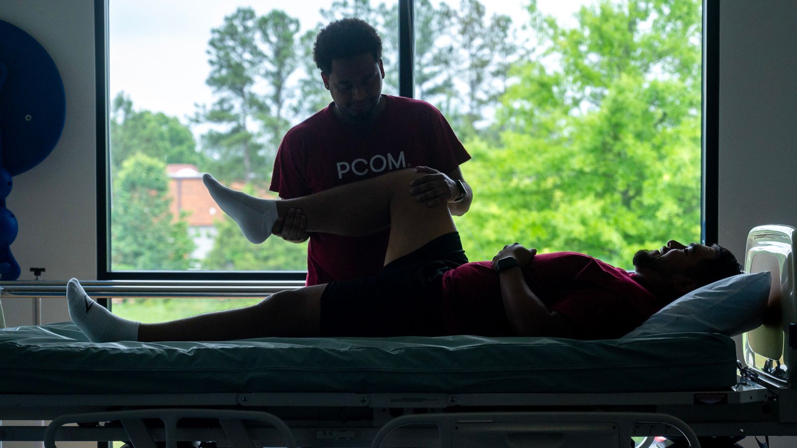 PCOM student in a maroon shirt performs a knee examination on a classmate lying on an exam table silhouetted against a window