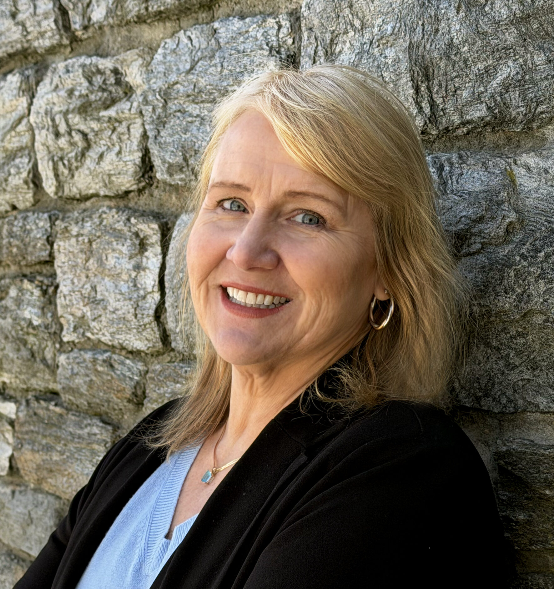 Abby Harmon smiles wearing a black sweater and blue blouse while standing near a stone wall.