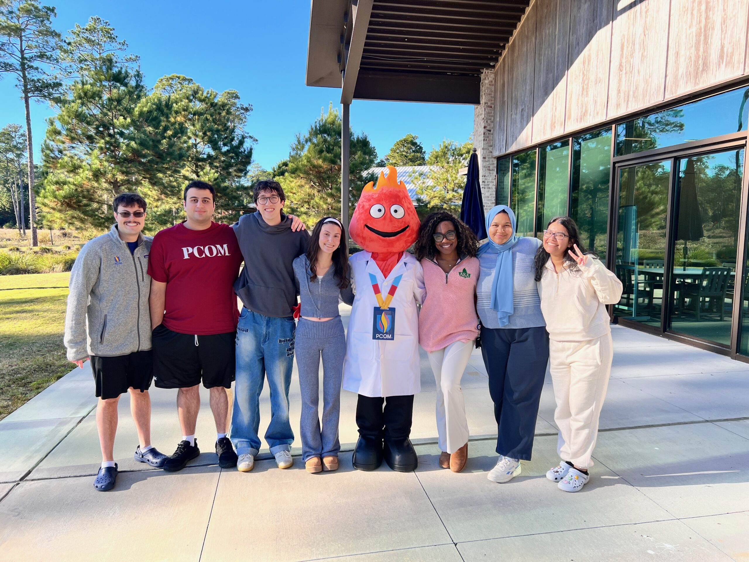 A mascot dressed in a flame head with white lab coat stands outside surrounded by a group of students.